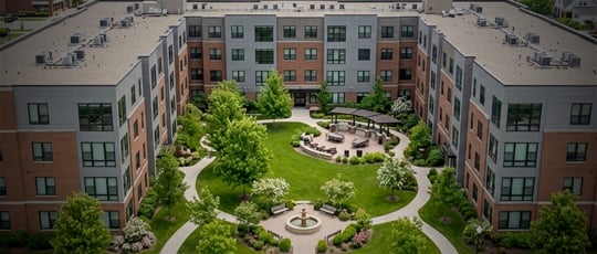 An aerial view of a multi-family residential apartment complex surrounding a landscaped courtyard with green grass, trees, and walking paths.